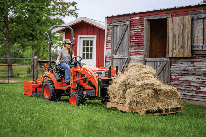 Woman using the Kubota subcompact tractor to move a pallet of hay